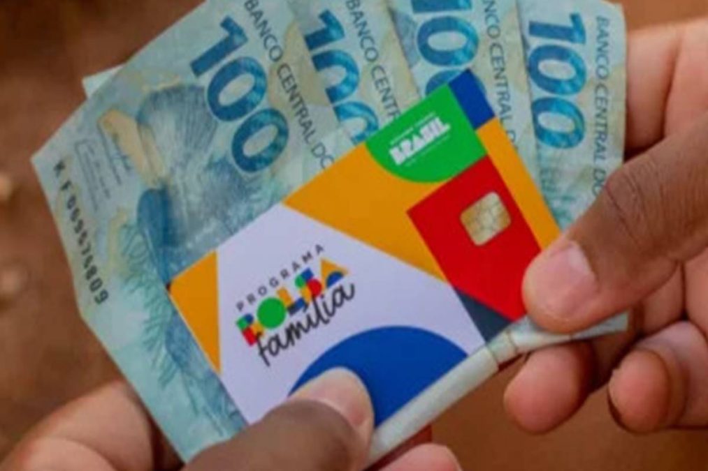 Pessoa segura cartão do Bolsa Família e notas de 100 reais